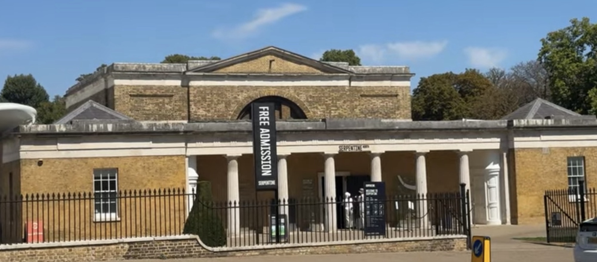 Yellow brick Serpentine gallery building with a Free Admission banner hanging from its columned entrance