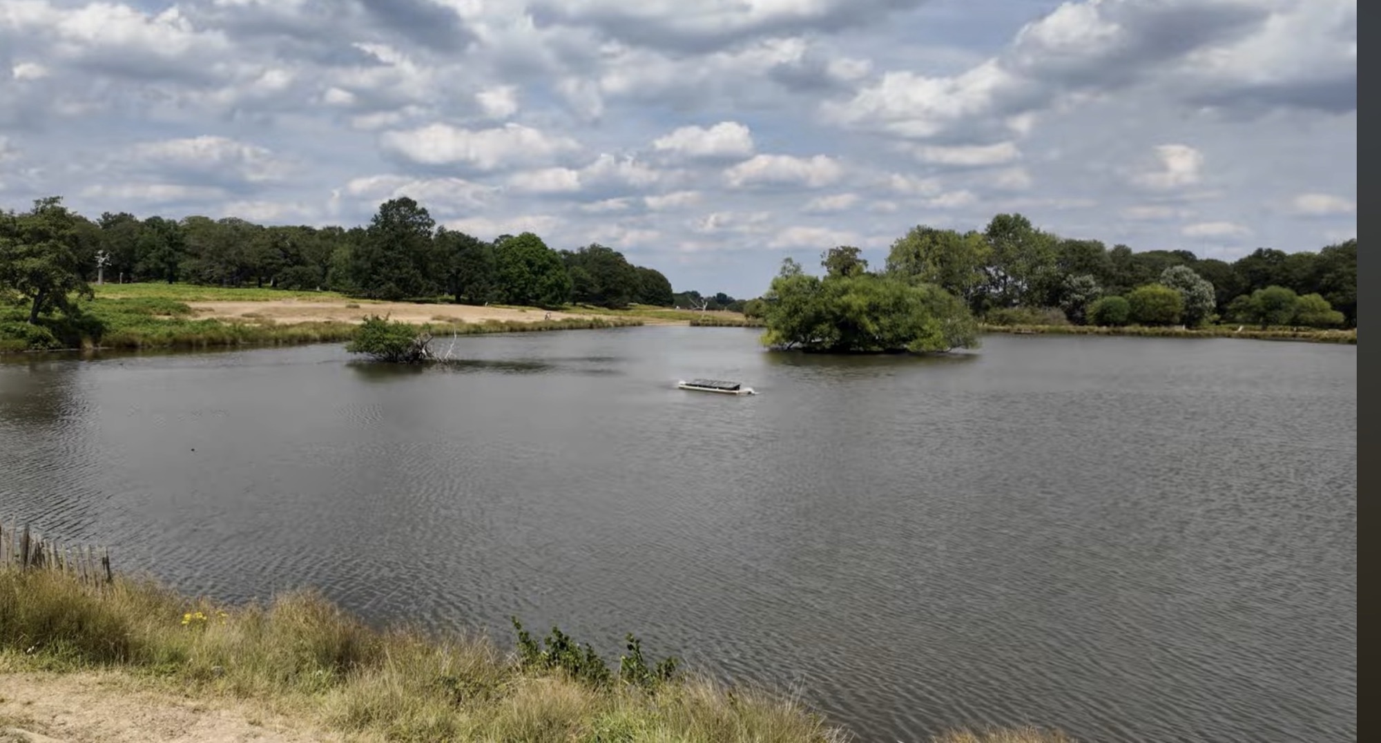 A wooded island and a moored pedalo on one of the Pen Ponds in Richmond Park under a cloudy sky