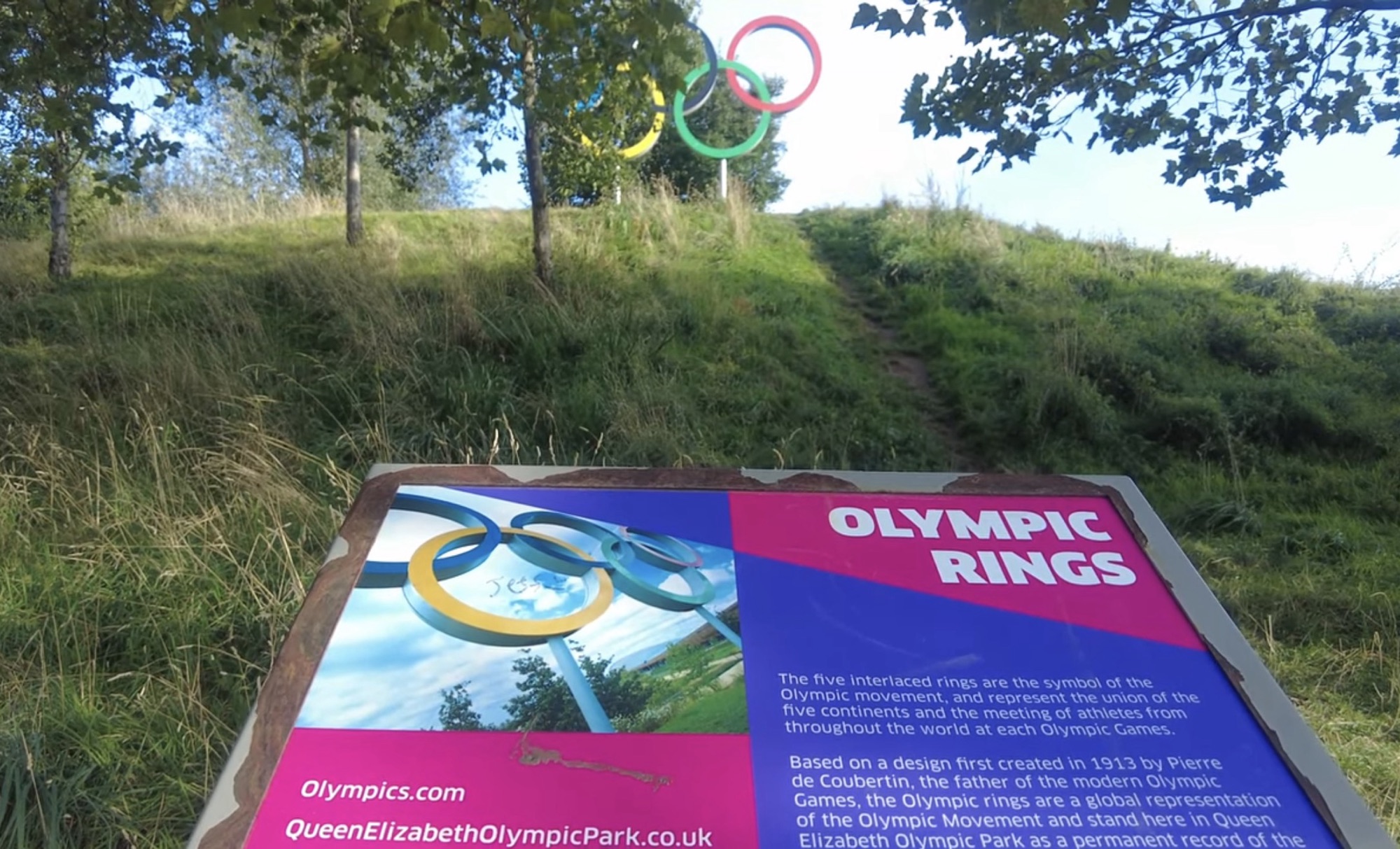 Olympic Rings sculpture on a grassy hill with an information board explaining their symbolism in Queen Elizabeth Olympic Park