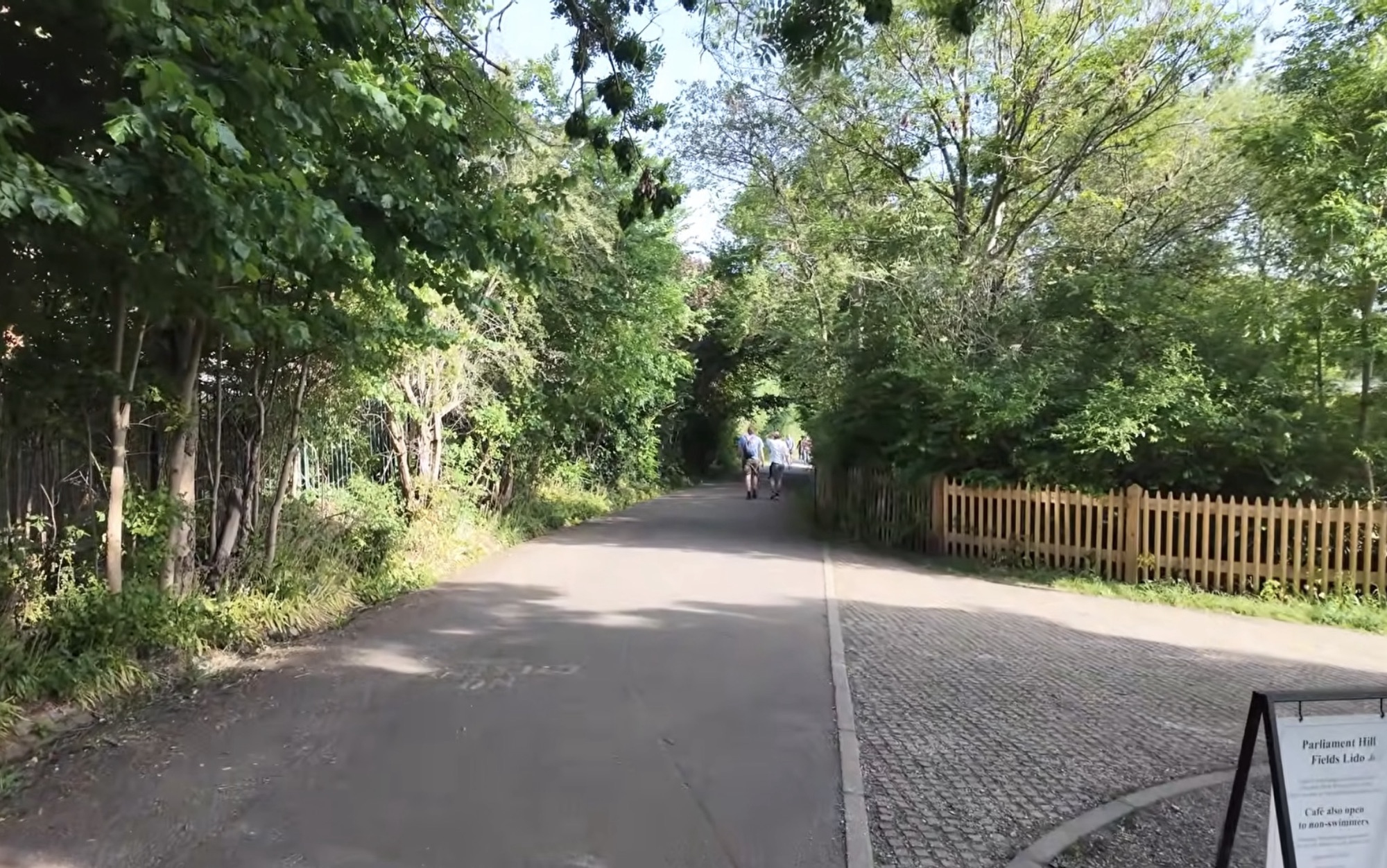 Tarmac path leading past a wooden picket fence towards Parliament Hill Fields Lido at Hampstead Heath