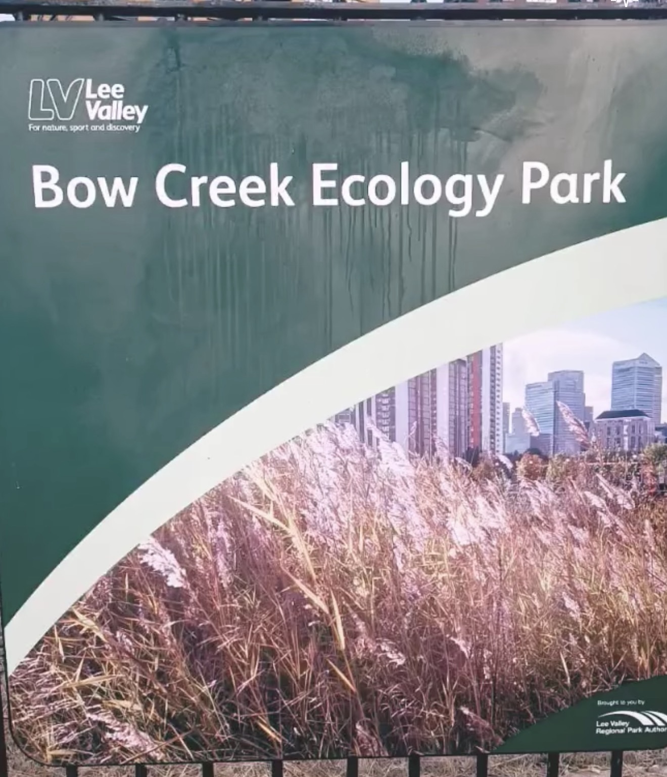 Bow Creek Ecology Park sign with reed beds and the Canary Wharf skyline behind