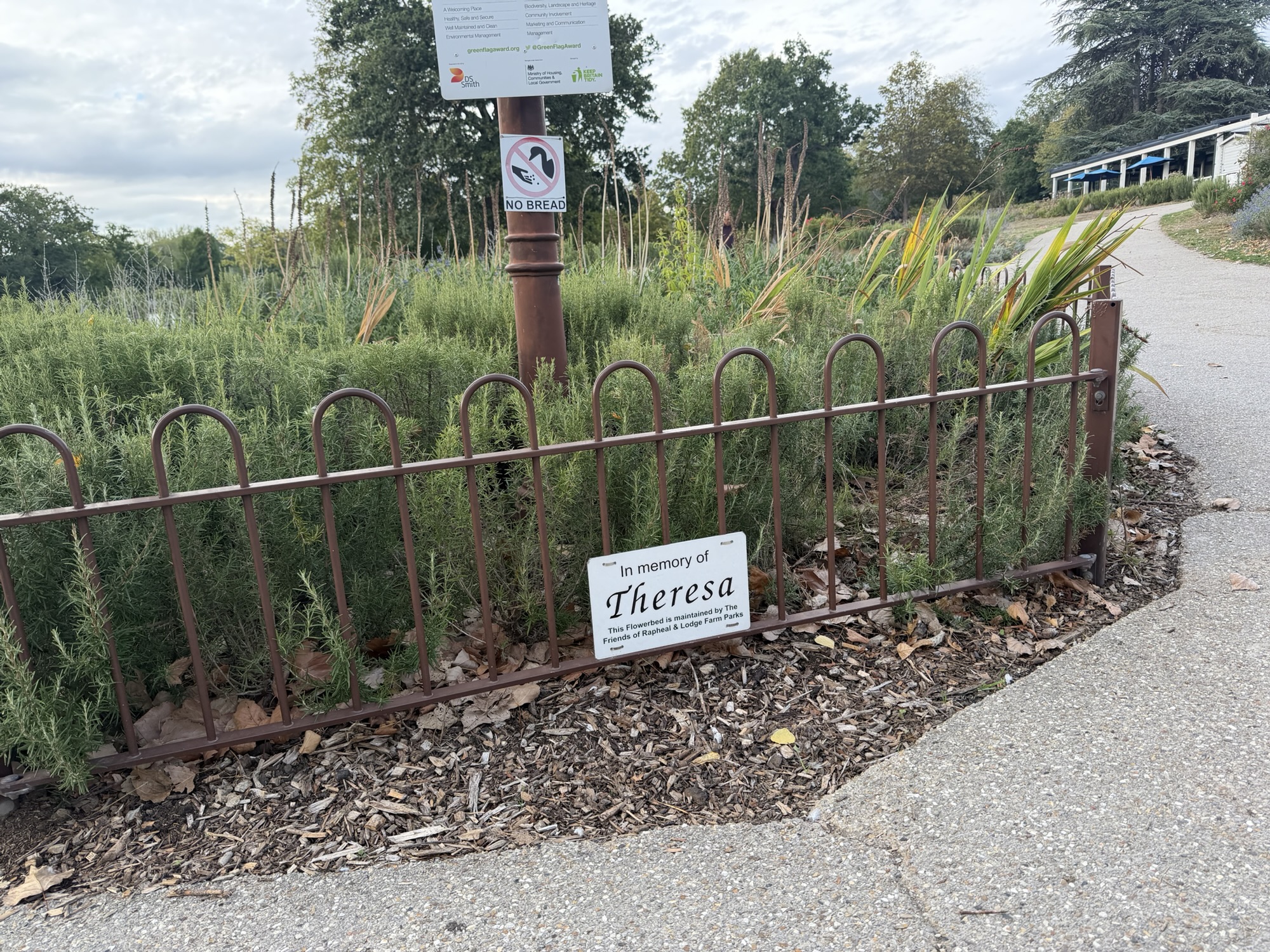Memorial plaque on a flowerbed fence reading In memory of Theresa, maintained by the Friends of Raphael and Lodge Farm Parks, next to a no bread sign