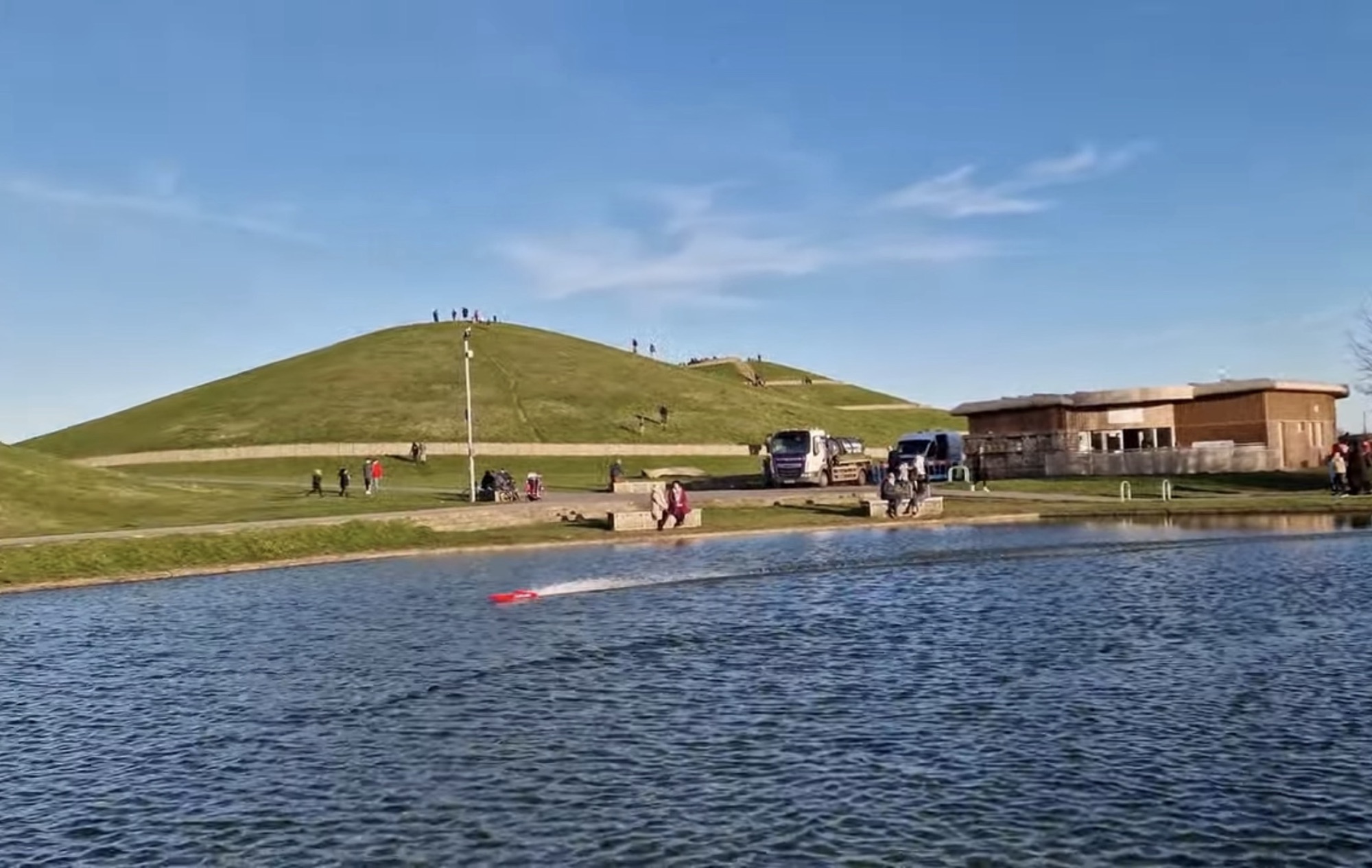Red model boat on the lake with a grassy mound and the visitor centre building behind at Northala Fields
