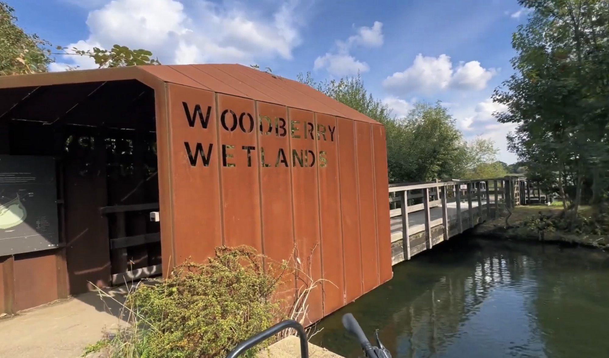 Rust-coloured steel entrance structure with Woodberry Wetlands cut into it, beside a wooden footbridge over the water
